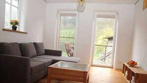 Un salon moderne avec un canapé gris, une table basse en bois et une table avec un plateau en verre. Il y a deux grandes fenêtres donnant sur un jardin, et un banc en bois à l’extérieur.