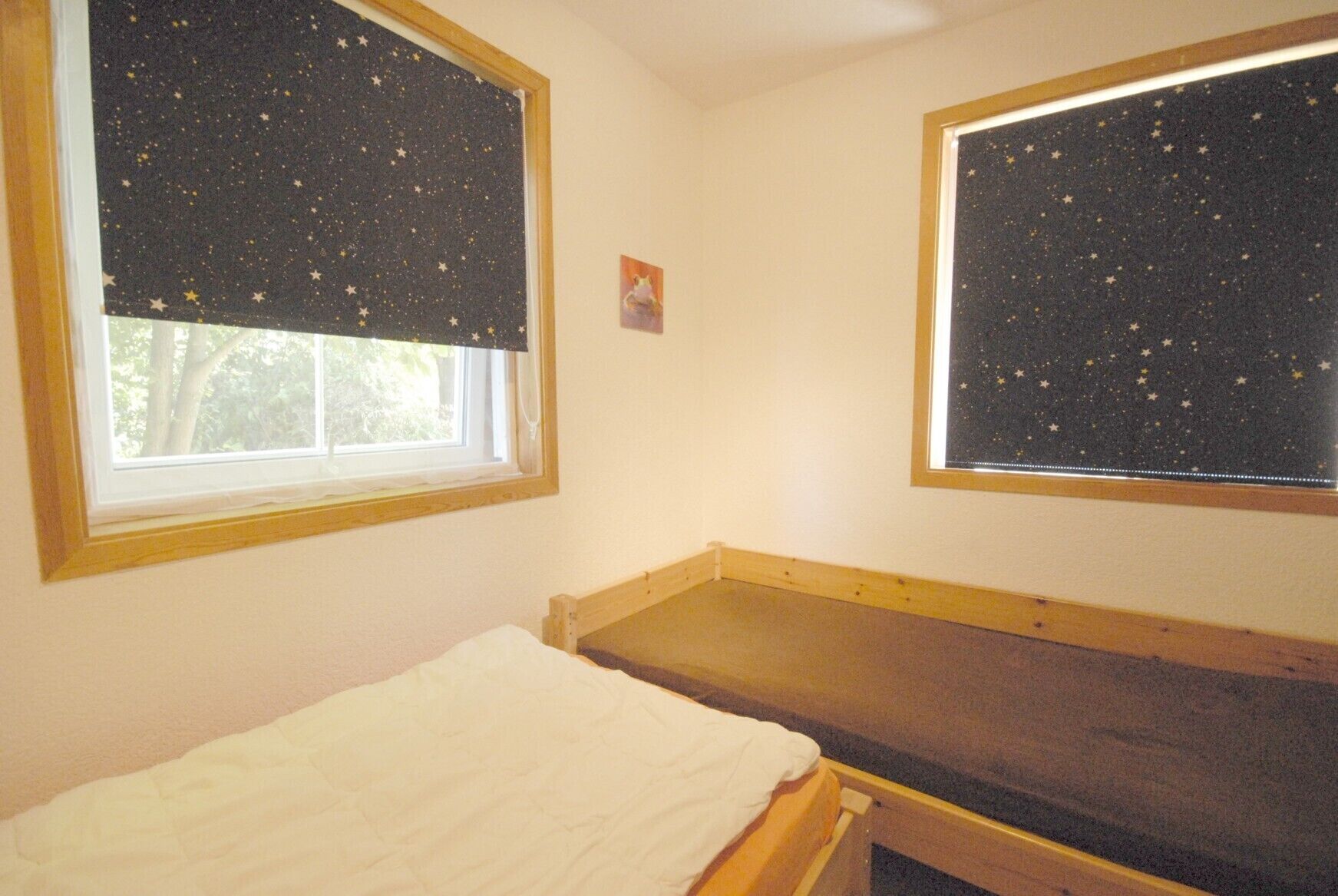 A small, simple room with a single bed, two windows with star-patterned curtains, and a small picture on the wall.