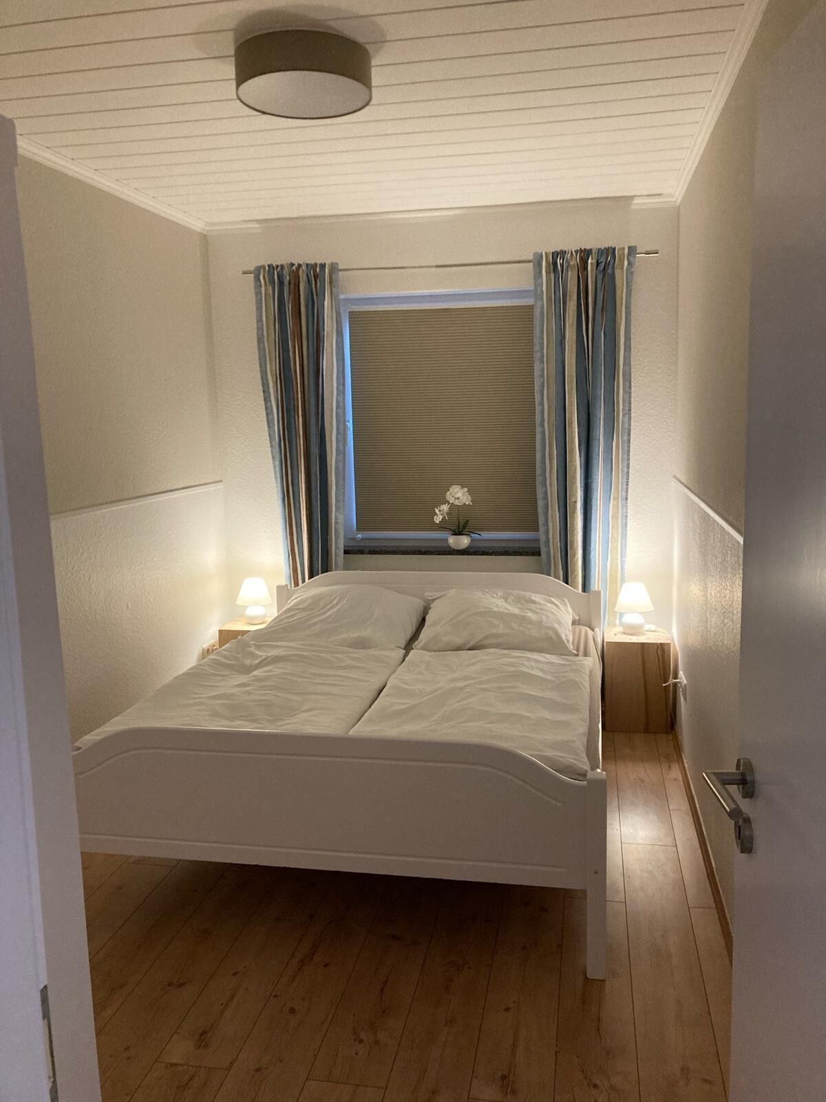 Una camera da letto con un letto, due lampade ai lati, una finestra con tende e una piccola pianta in vaso sul davanzale.