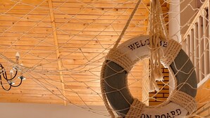 Una habitación acogedora con una hamaca con la leyenda “BIENVENIDOS A BORDO” colgando del techo, un sofá con cojines, una chimenea y vistas al exterior a través de un ventanal grande.