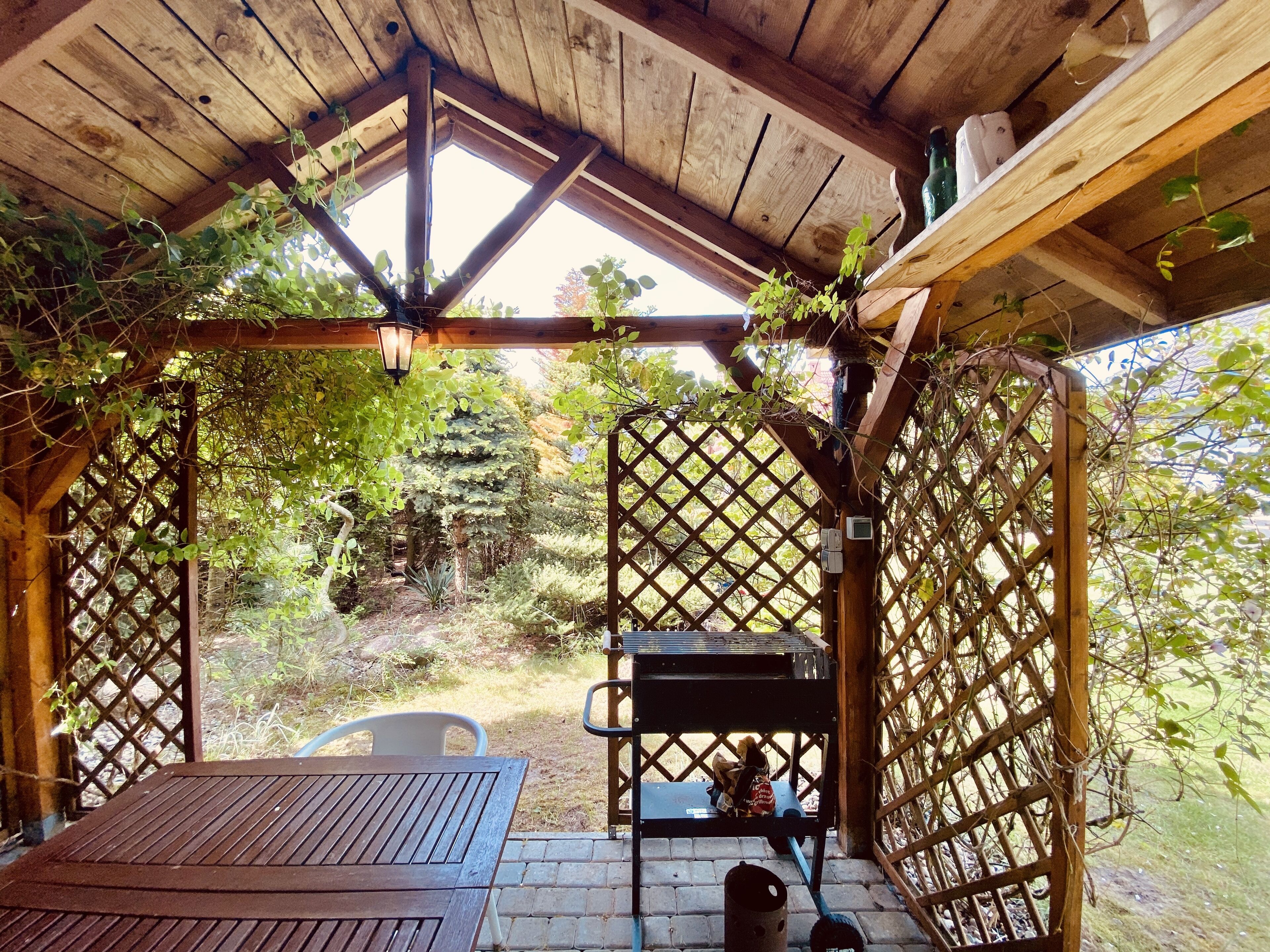 Uma pérgola de madeira com mesa e cadeiras, um candeeiro suspenso e vista para um jardim com árvores e um banco.