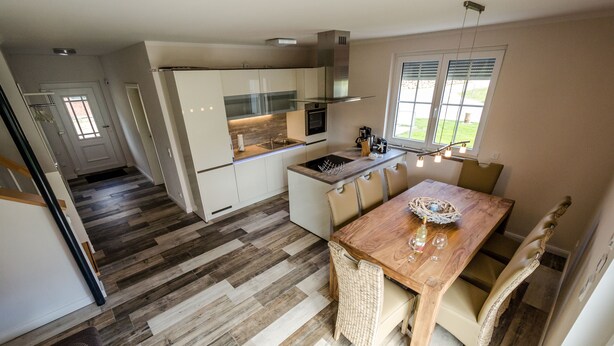 Une cuisine moderne dotée d’un îlot central, d’un coin repas avec une table et des chaises en bois, et offrant une vue sur l’extérieur grâce à de grandes fenêtres.