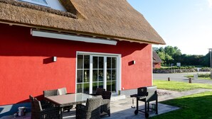 Un bâtiment rouge avec un toit de chaume, une terrasse meublée de meubles en osier et une pelouse bien entretenue.