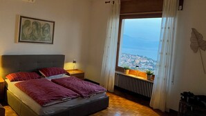 A bedroom with a bed, a nightstand, a lamp, a radiator, a window with curtains, and a view of a cityscape. - Romantic villa with a fantastic view of Lake Garda (Toscolano Maderno)