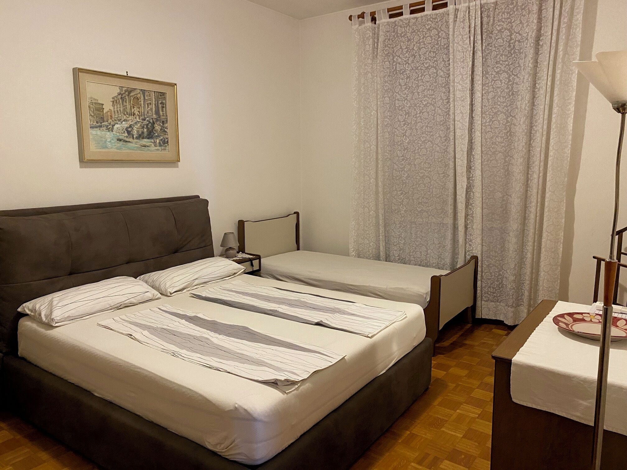 Ein Hotelzimmer mit einem großen Bett, einem kleineren Bett, einem Nachttisch, einer Lampe, einem Bild an der Wand und einem Fenster mit Vorhängen.