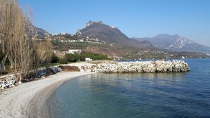 A coastal area with a pebble beach, a stone breakwater, and a backdrop of mountains and buildings. - Romantic villa with a fantastic view of Lake Garda (Toscolano Maderno)