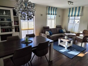 A living room with a dining table, sofa, and chairs, a bookshelf, and a large window with curtains.