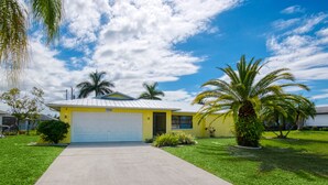 A yellow house with a white garage door, surrounded by well-maintained grass and palm trees. - Villa Sunsplash-Modern, south-facing, large pool- perfect for families (Cape Coral)