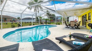 A covered outdoor swimming pool with lounge chairs and a table, surrounded by a white fence and palm trees. - Villa Sunsplash-Modern, south-facing, large pool- perfect for families (Cape Coral)