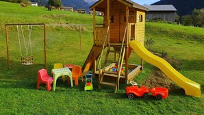 Une maison de jeu en bois, avec un toboggan et des balançoires, située dans un champ verdoyant, avec des montagnes en arrière-plan.