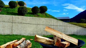Un foyer en métal noir, avec des bûches, installé sur de l’herbe, avec un mur en béton et des collines en arrière-plan.