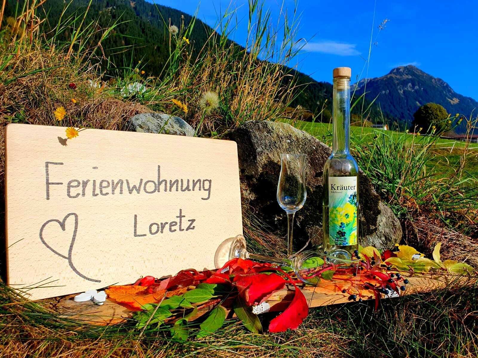 A wooden sign with a heart and the text "Ferienwohnung Loretz" in a natural outdoor setting with a bottle of Kräuter and a glass.