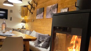 A cozy room with a wood-burning stove, a dining table with chairs, a sofa, and a wall-mounted TV.