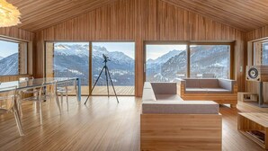 A modern interior with wooden paneling, a glass table with chairs, a tripod, and a sofa with a mountain view. - Exclusive design chalet on 1 800 meters, 4-6 pers.,  above Törbel (Törbel)