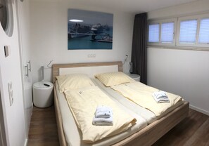 A double bed with two pillows, a bedside table, a lamp, a waste bin, and a painting of a harbor scene. - Exclusive apartment in Warnemünde, 150m to the beach, 2 bathrooms, 2 bedrooms, sauna, WLAN and much more. (Rostock)