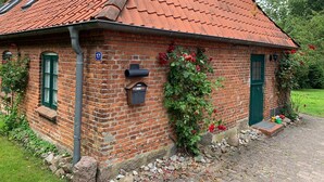 A brick house with a red-tiled roof, a chimney, and a small garden with red flowers. - Idyllic farm cottage only 600 m from the Baltic Sea, natural beach, no car traffic (Barkelsby-Hemmelmark)