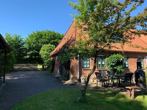 A brick house with a red-tiled roof, a garden with a table and chairs, and a wooden fence in the background.