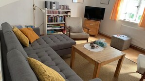 A living room with a grey sectional sofa, a wooden coffee table, a bookshelf, a television, and a window with curtains. - Idyllic farm cottage only 600 m from the Baltic Sea, natural beach, no car traffic (Barkelsby-Hemmelmark)