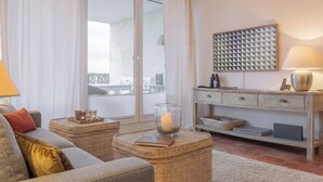 Un salon moderne comprenant un canapé, deux poufs en osier, un porte-bougie en verre, un bureau avec des tiroirs, une lampe et un diffuseur sonore fixé au mur.