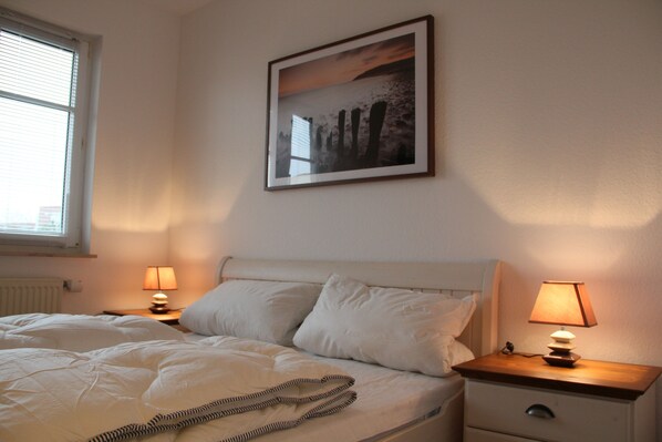 A neatly made bed with white linens and pillows, two bedside lamps, a framed picture on the wall, and a window with blinds.