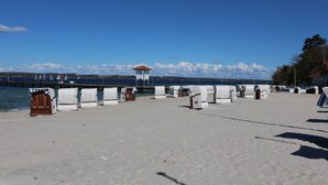 Een strand met witte strandstoelen en een houten bank, een redershut en een pier met zeilboten op de achtergrond.
