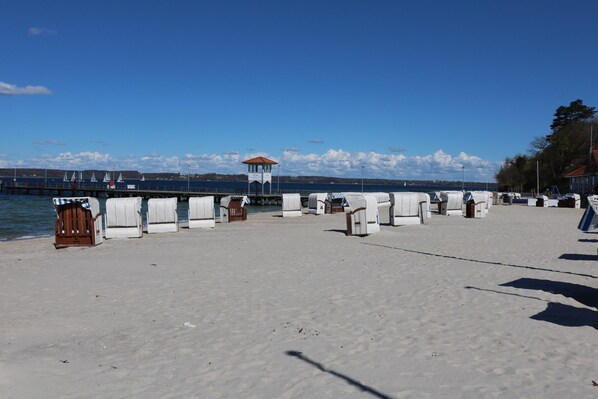 Een strand met witte strandstoelen en een houten bank, een redershut en een pier met zeilboten op de achtergrond.