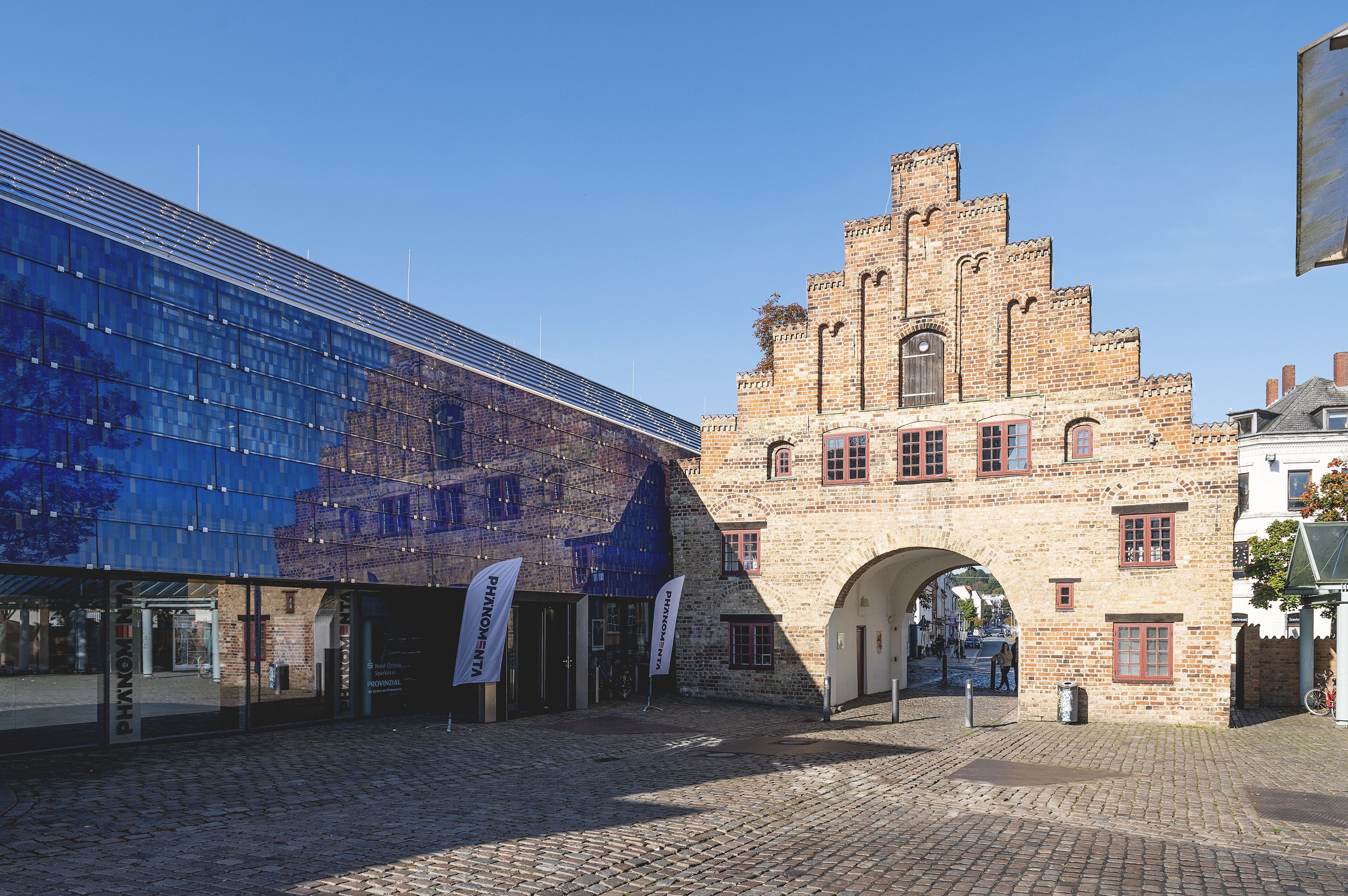 A cobblestone street with a historic building featuring a tower and a modern glass structure.