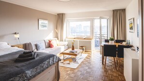 Een hotelkamer met een bed, een bank, een eettafel met stoelen, een raam met gordijnen en uitzicht op de stad.