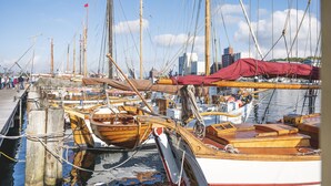 Een jachthaven met verschillende houten boten afgemeerd, waarvan sommige afgedekt zijn met zeilen, en een stadsgezicht op de achtergrond.