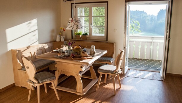 Een houten eettafel met stoelen, een bank en een raam met uitzicht op groen.