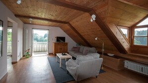 Ruime loft met schuine kap, een bank, een bijzettafeltje, een televisie op een houten kast, en uitzicht op groen.