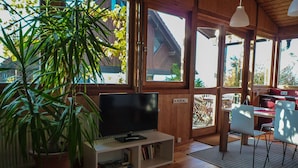 A wooden interior with a television on a stand, a potted plant, and a view of the outside through large windows. - Pure relaxation! Holiday home: integrated into the nature with modern interior! (Frickingen-Altheim)