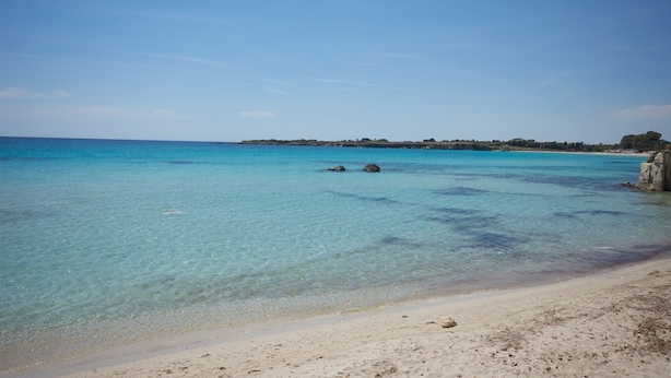 Una playa con agua turquesa cristalina, orilla de arena y costa rocosa en la distancia.