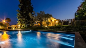 Una piscina con superficie de agua azul cristalina, rodeada de vegetación cuidada y una casa con iluminación exterior.