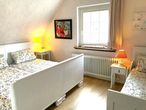 Ein Schlafzimmer mit zwei Betten, einem Fenster mit Vorhängen, einem Heizkörper und einem Nachttisch mit Lampe.