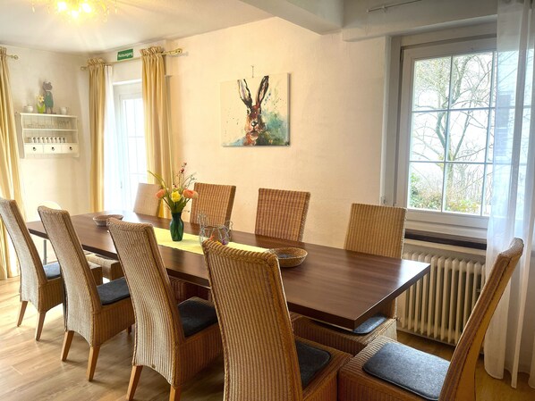 A dining area with a wooden table, wicker chairs, a vase with flowers, and a painting on the wall. - Family-friendly home for 10 persons, big garden, Fireplace, 4 km ski carousel. (Winterberg)