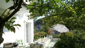 An outdoor dining area with a long table set for a meal, surrounded by greenery and a white building.