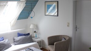 Ein gemütliches Zimmer mit schrägen Wänden, einem Bett mit blauen Kissen, einem kleinen Tisch mit einer Lampe und einem Sessel.
