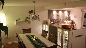 Ein Essbereich mit einem Tisch für vier Personen, umgeben von Korbstühlen. Im Hintergrund ist eine Küche mit Arbeitsplatte und Schränken erkennbar.