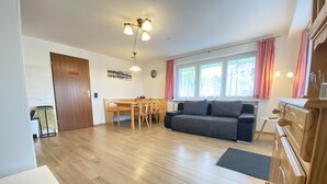 Ein modernes Wohnzimmer mit Holzboden, einer Couch, einem Esstisch mit Stühlen, einer Küchenzeile mit Herd und einem Fenster mit Vorhängen.