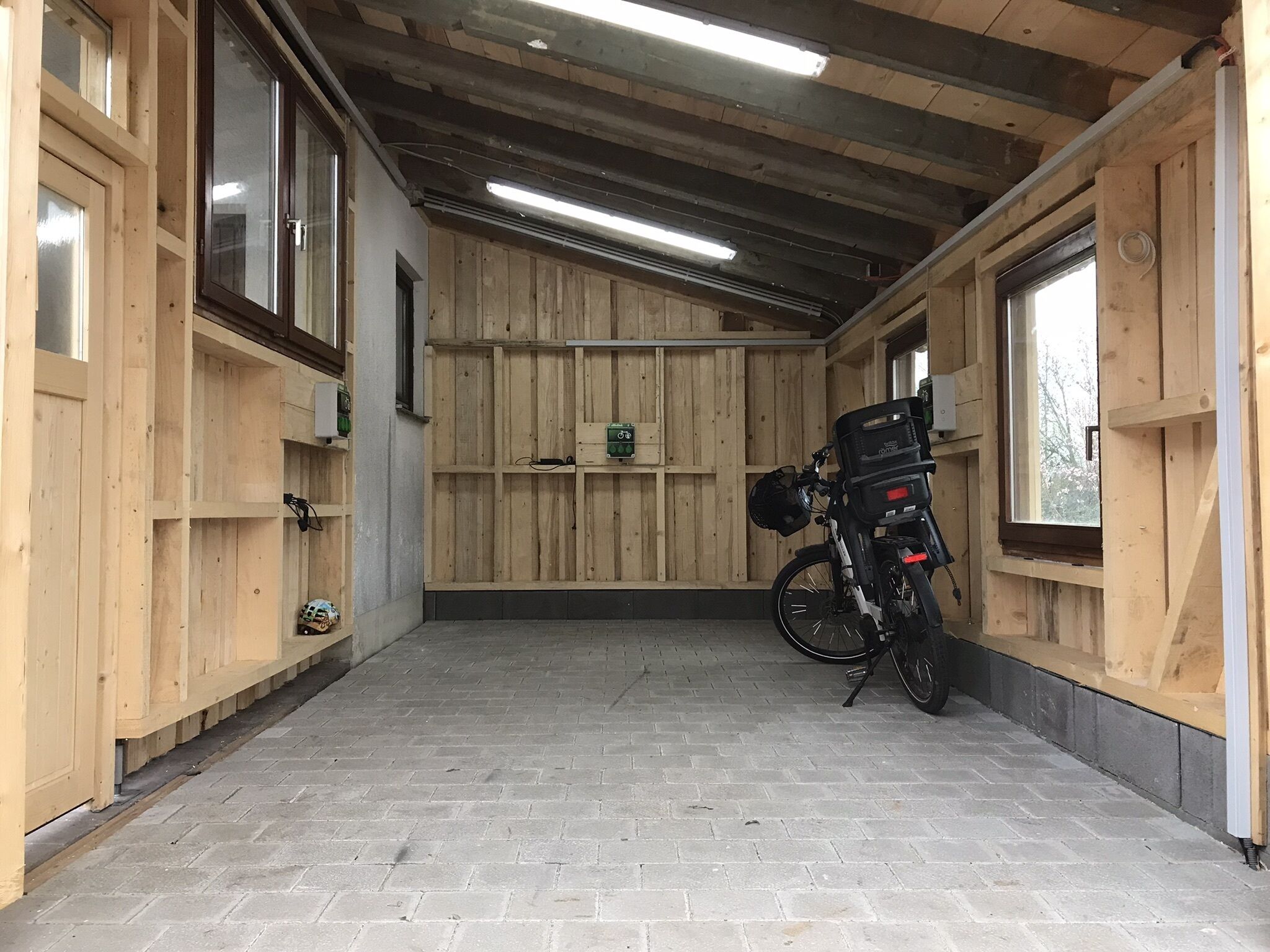 A covered bike parking area with a wooden floor and walls, a bicycle with a basket, and a window on the right.