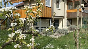 Ein modernes, zweistöckiges Haus mit Holzfassade, einem sichtbaren Balkon und einem Garten mit blühendem weißen Flor.