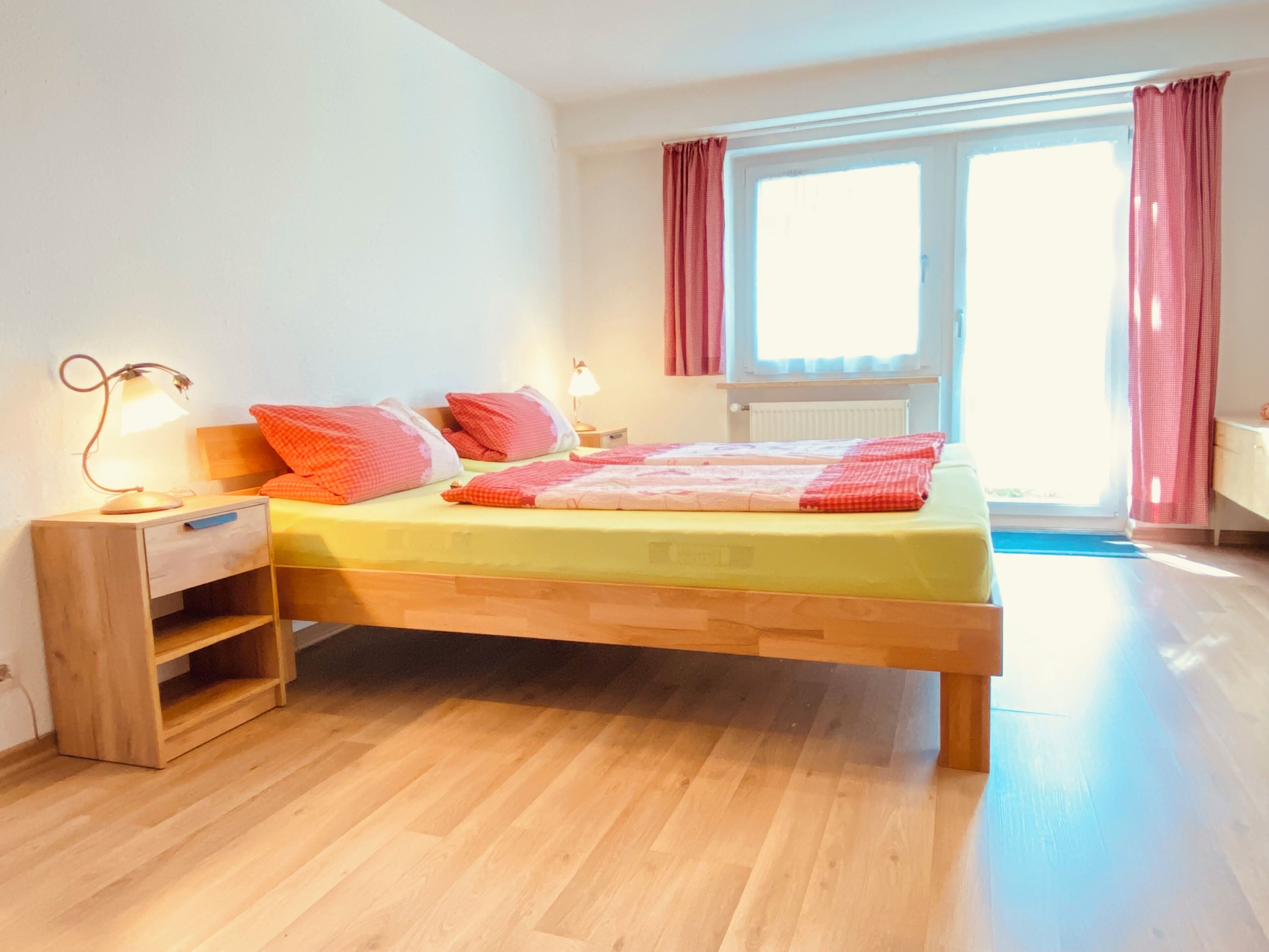 A bedroom with a wooden bed, a nightstand with a lamp, a window with curtains, and a view of the outdoors.