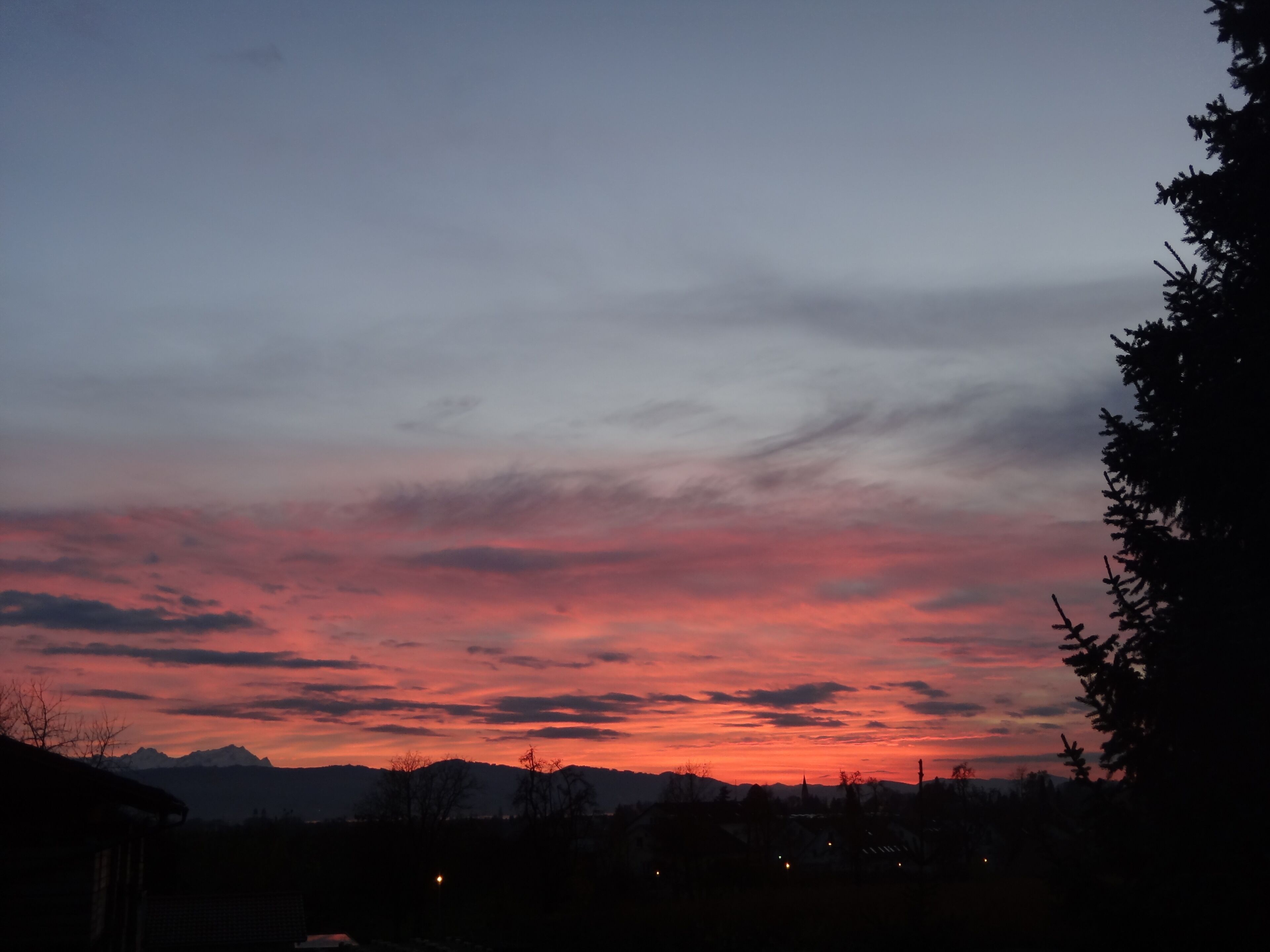 Auringonlasku vuoristomaisemassa, jossa näkyy puiden ja rakennusten siluetteja.