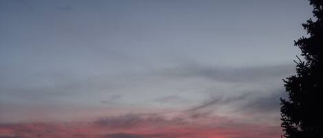 Auringonlasku vuoristomaisemassa, jossa näkyy puiden ja rakennusten siluetteja.