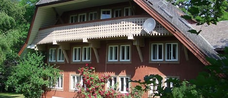 A two-story house with a balcony, surrounded by greenery and a bench.