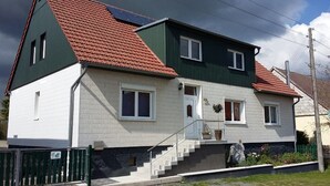 A two-story house with a red-tiled roof, white walls, and a green exterior. It features a front entrance with a staircase, a small garden, and a solar panel on the roof. - "Pure nature" ... Vacation in the Harz Mountains (Stiege)