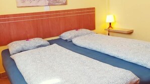 Een tweepersoonsbed met een blauwe matras en witte beddengoed met een patroon, een houten hoofdbord, een nachtkastje met een lamp en een schilderij aan de muur.