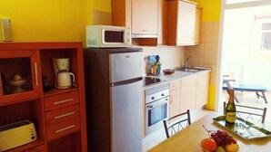 Een kleine keuken met een magnetron, koelkast en kookplaat. Er is een eettafel met een mand fruit en een fles wijn.