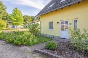 Um edifício amarelo com porta e janelas brancas, rodeado por um jardim com diversas plantas e um caminho pavimentado.